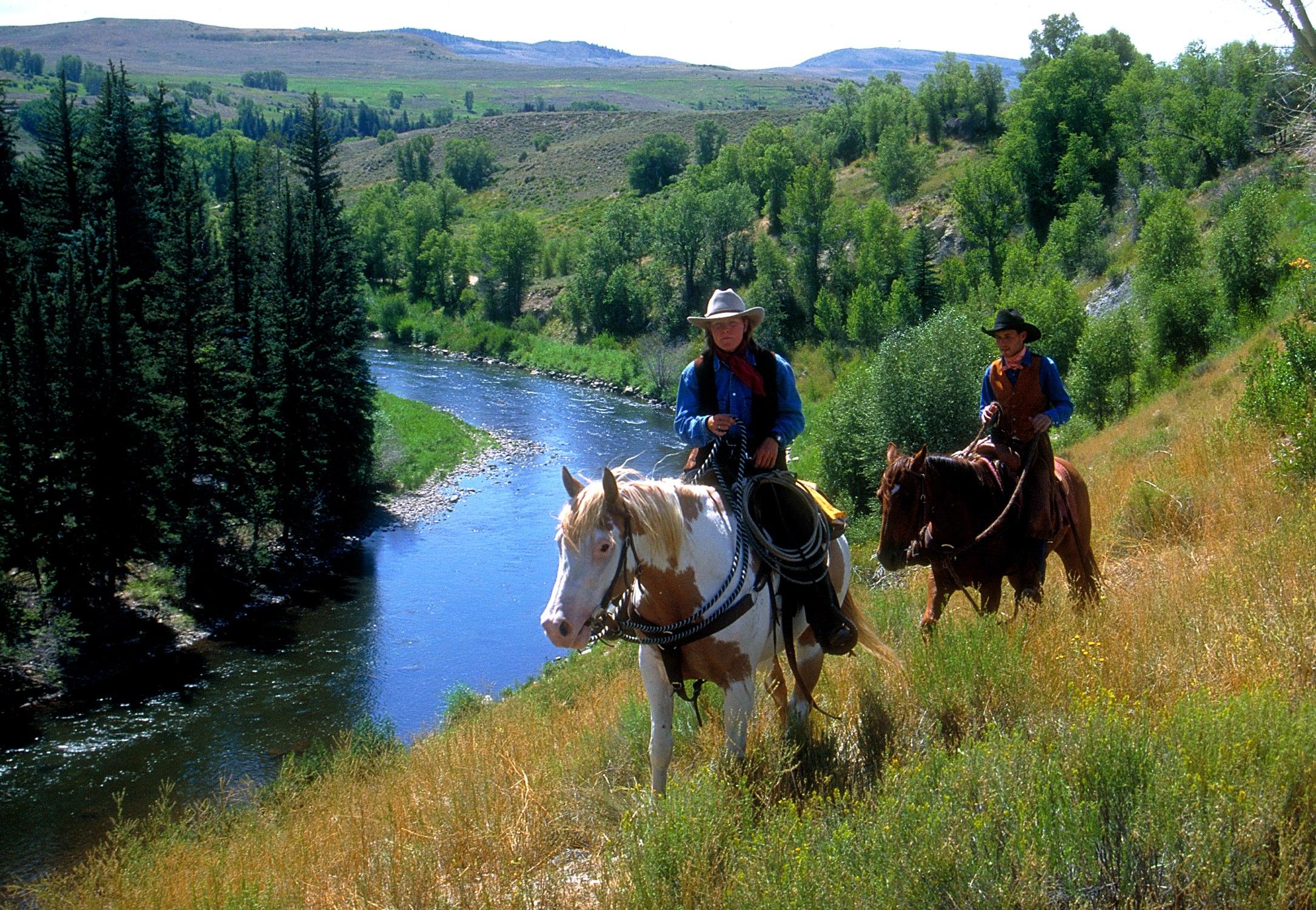 Two people dressed as cowpokes each ride a horse through a hillside field. Beside them flows a small body of water.