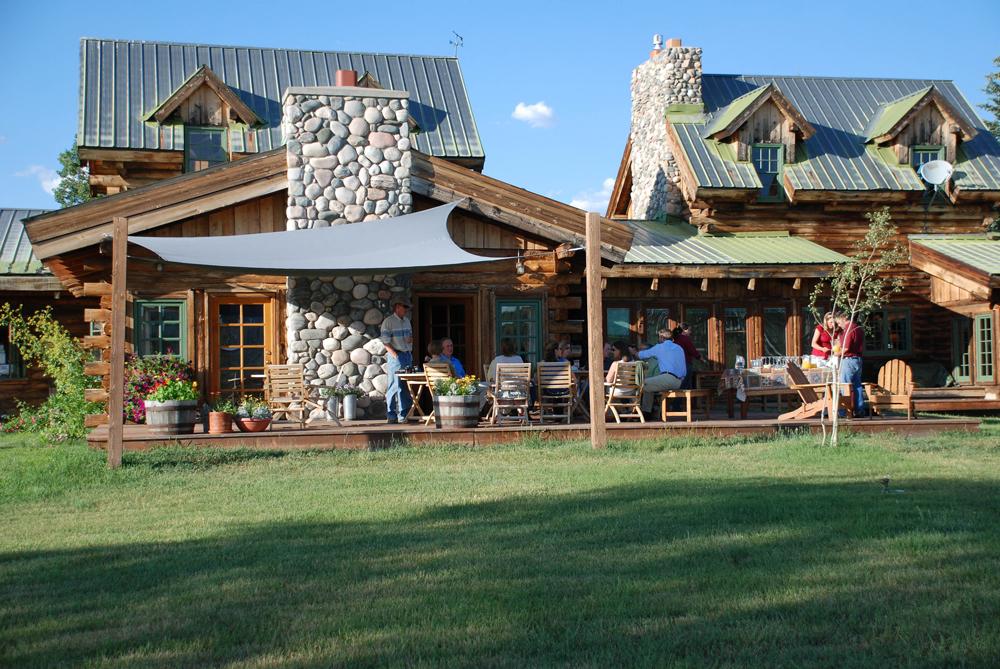 A ranch lodge with a stone chimney and expansive front porch sits amid a green meadow. Guests relax on and children play in front of them