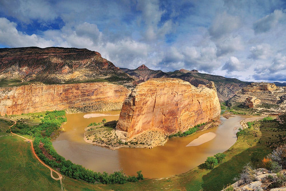 Multi-layered rock plateaus sit in the background under a blue, cloudy sky. In the middle there's a red rock surrounded by the confluence of the Yampa and Green Rivers. It's summer and the riverbanks are covered in green grass and scrubby green trees.