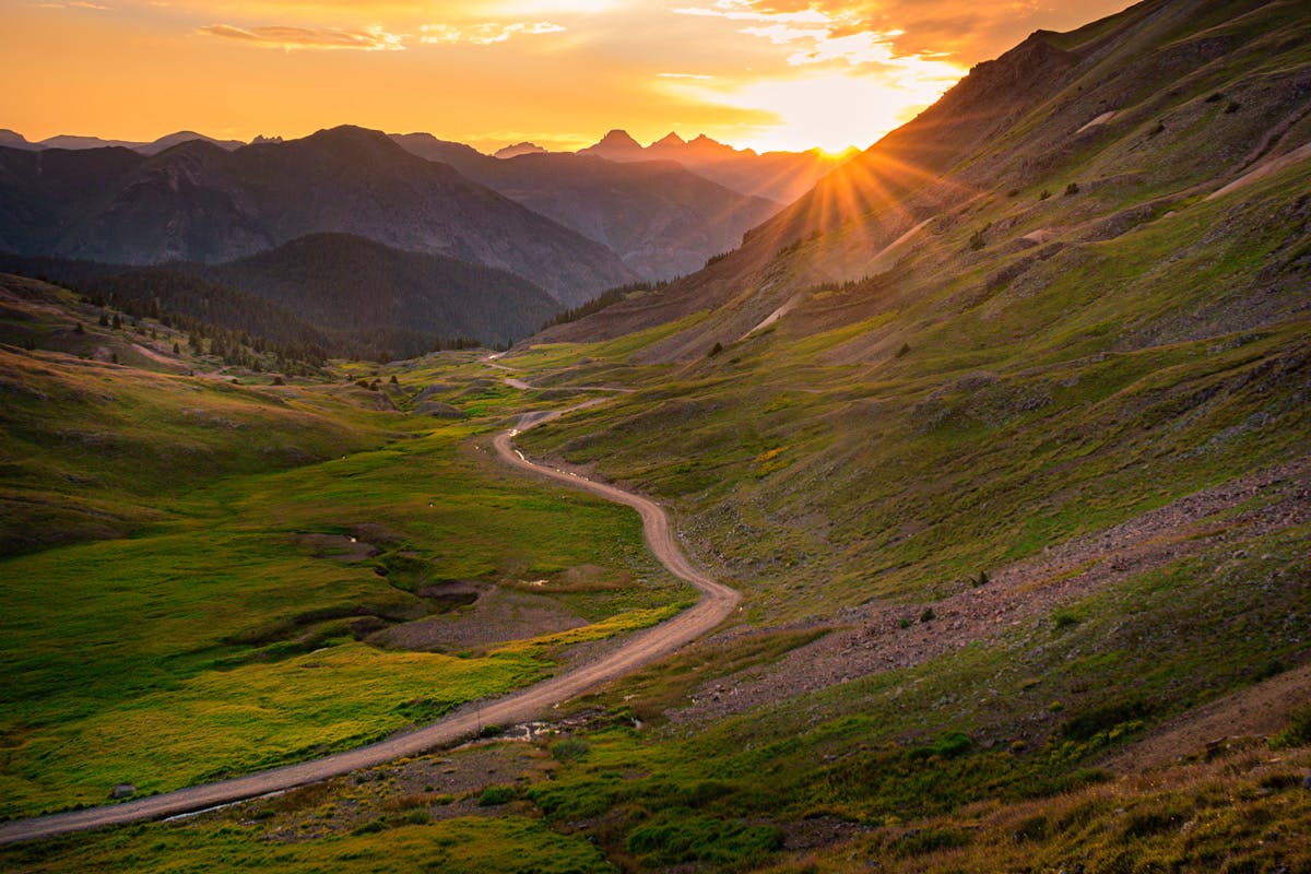 As the sun sets behind mountain peaks, an off-road path with green grasses sits in the middle of the image.