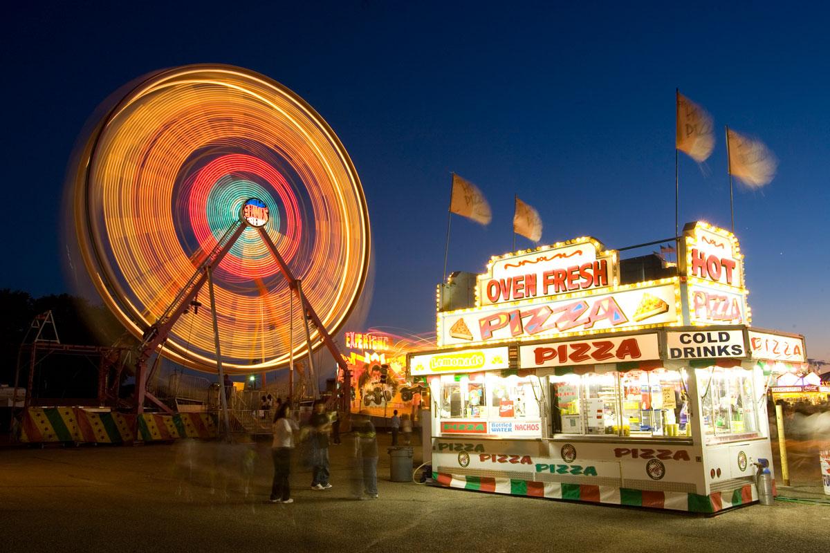 The yellow, red and lights of a Ferris wheel and the white lights of a food stand sit beneath a darkening sky