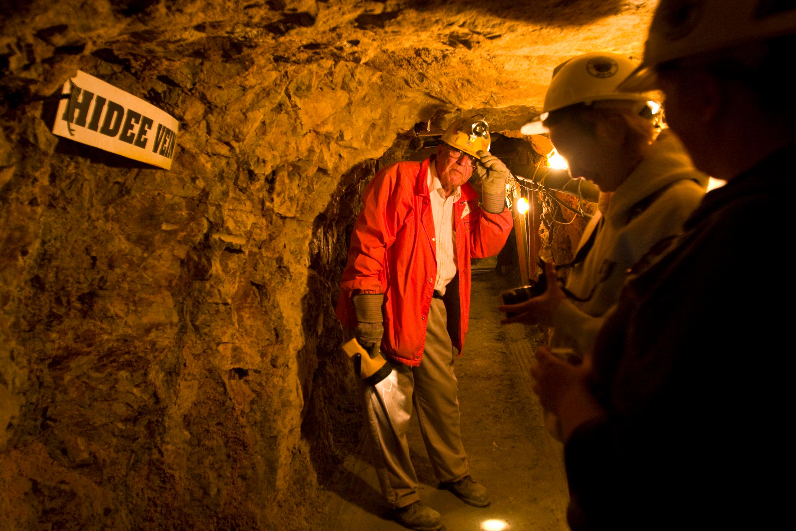 A tour guide in a red jacket and yellow hard hat leads a group of people through a dimly lit tunnel