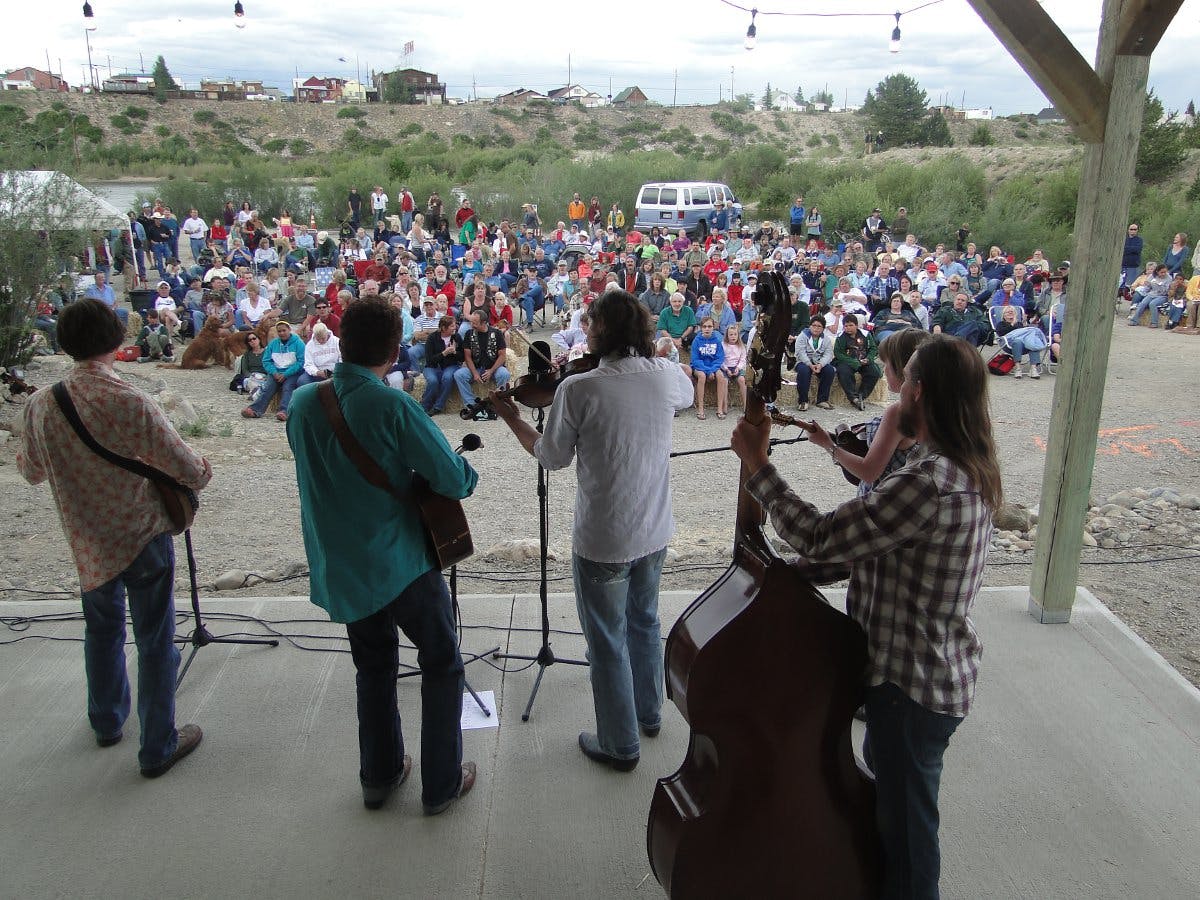 Four people facing a large crowd play an acoustic concert in FairPlay. In the distance scrubby greenery sits atop grey-tan dirt and town is in the distance.