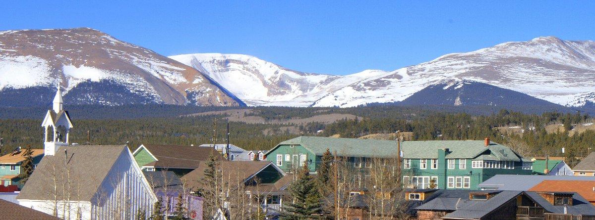 Snow-covered mountains meet a bright-blue sky in the distance behind Fairplay's historic downtown. There are evergreen trees.