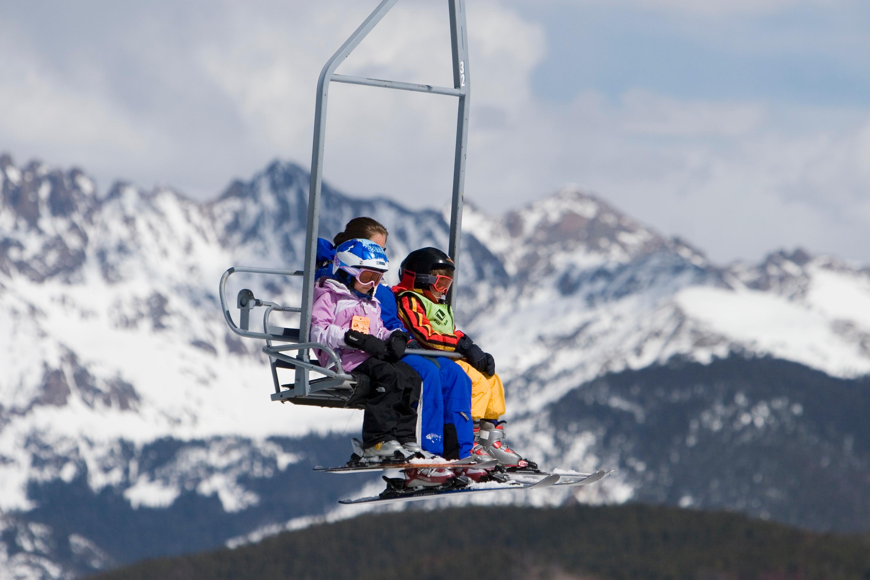 A mom and two kids ride a chairlift with all their ski gear. Behind them, we see snowy peaks.