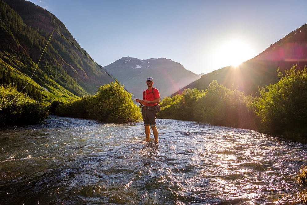 A man in a red t-shirt stands in the middle of a river stream smiling at the camera. Surrounded by green vegetation and in the distance there are mountain peaks and a blue sky with the setting sun.