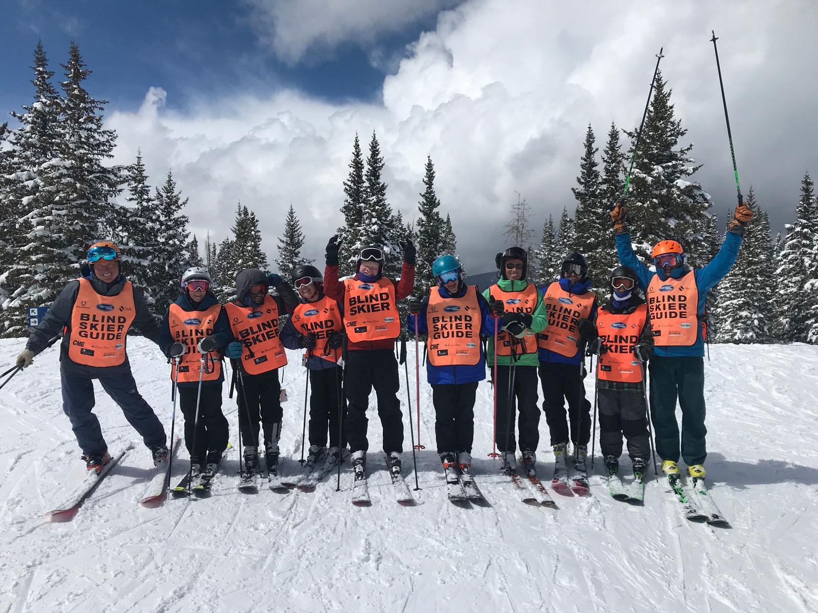 A group of skiers wears bright-orange bibs saying "Blind Skier" or "Blind Skier Guide" on a mountain slope in Colorado.