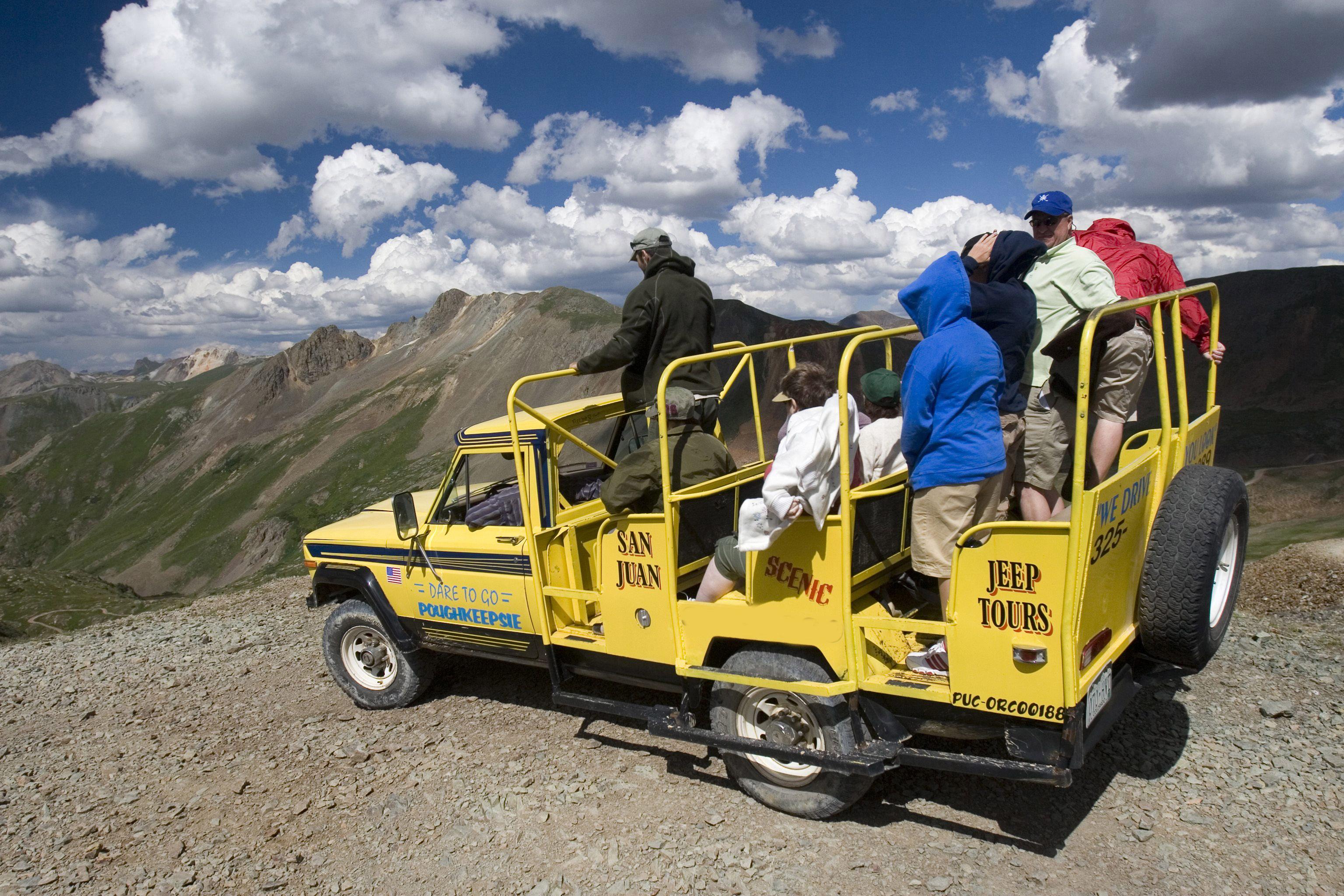 A yellow open-air tour bus sits on a rocky viewpoint with people looking out at Rocky Mountain tops with a blue sky and white clouds.