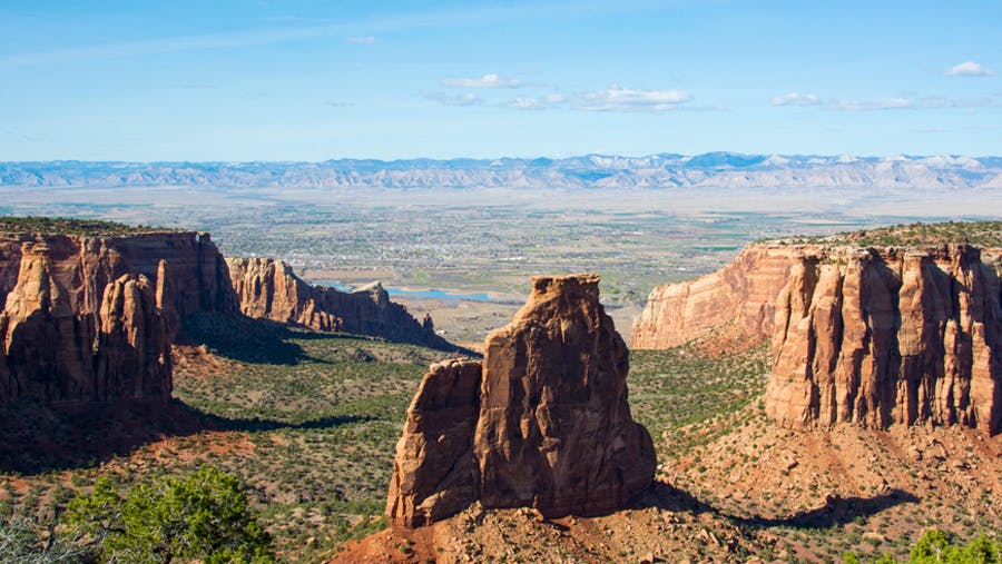 Three red-rock formations soar out of the green, scrubby Earth at Colorado National Monument. In the distance is a hazy, rocky ridge that meets a light-blue sky.