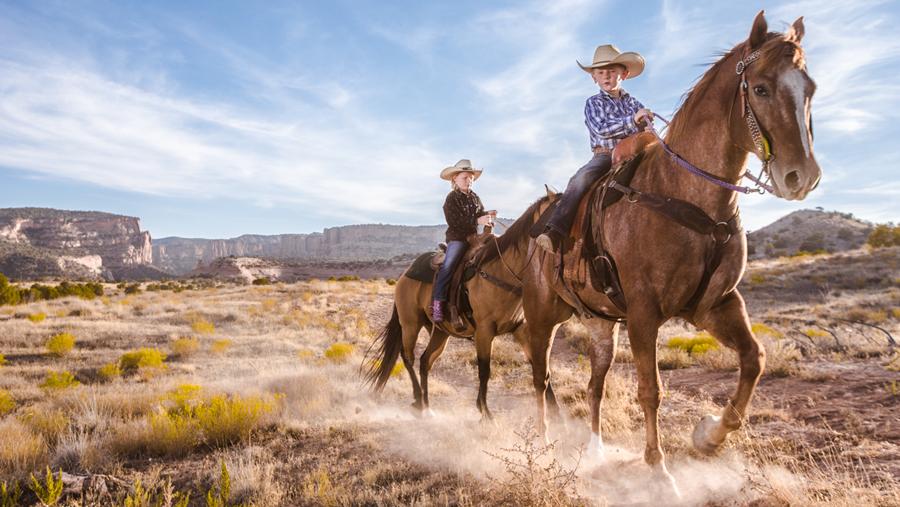 Two kids horseback ride down a sandy path, through brown and yellow scrubby grass under a blue sky with whispy white clouds. In the distance sheer rock walls emanate from the ground. 