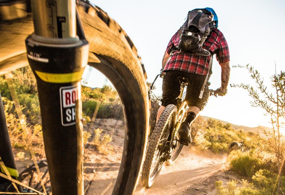 A bike wheel close-up with a mountain biker wearing a red shirt and backpack in the background make their way down a dirt singletrack at golden hour. There are green scrubby bushes on both sides.