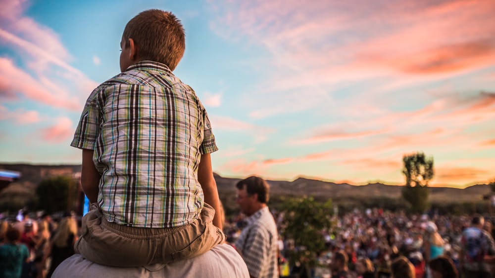 A kid wearing a plaid shirt sits on a person's shoulders at a sunset concert in Fruita, Colorado. On the right is an amassed crowd under a pink-cloud, light-blue sky.