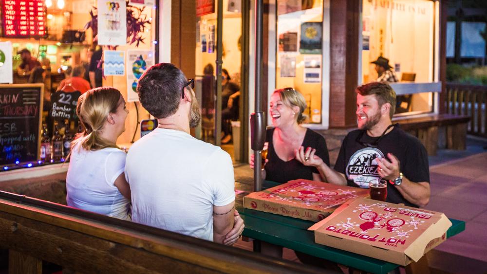 Two couples sit at a green picnic table on a sidewalk in Fruita. There are two cardboard pizza boxes on the table. In the background, a storefront with lots of windows glows.