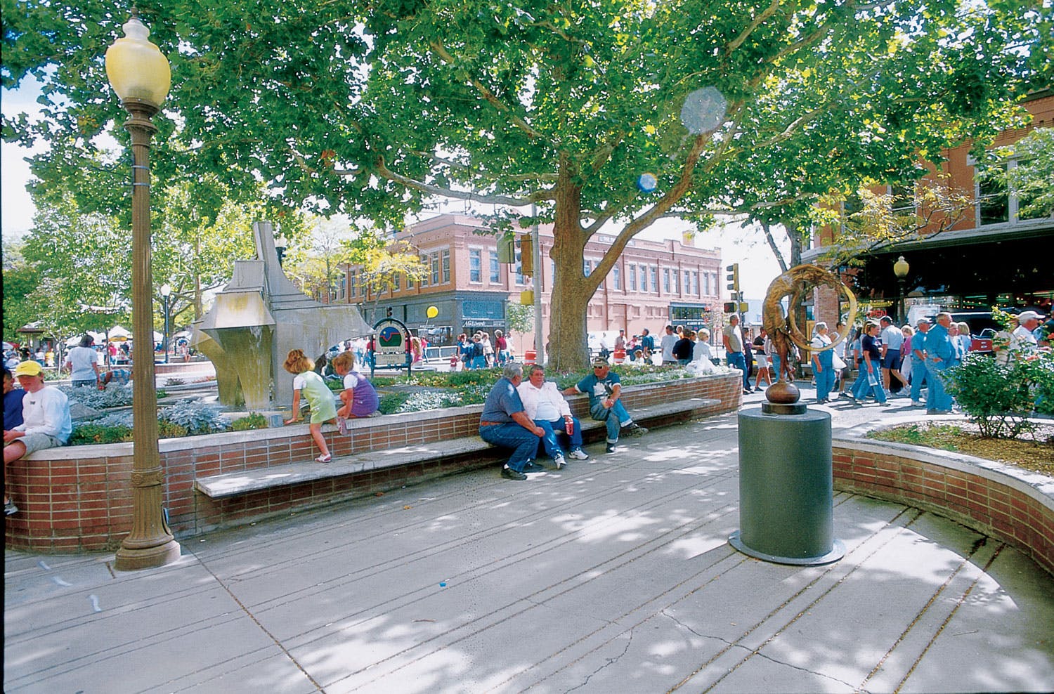 Lots of people sit in a a public park in downtown Grand Junction with green-leafed trees, low brick walls and public art. In the background there are two-story red-brick buildings.