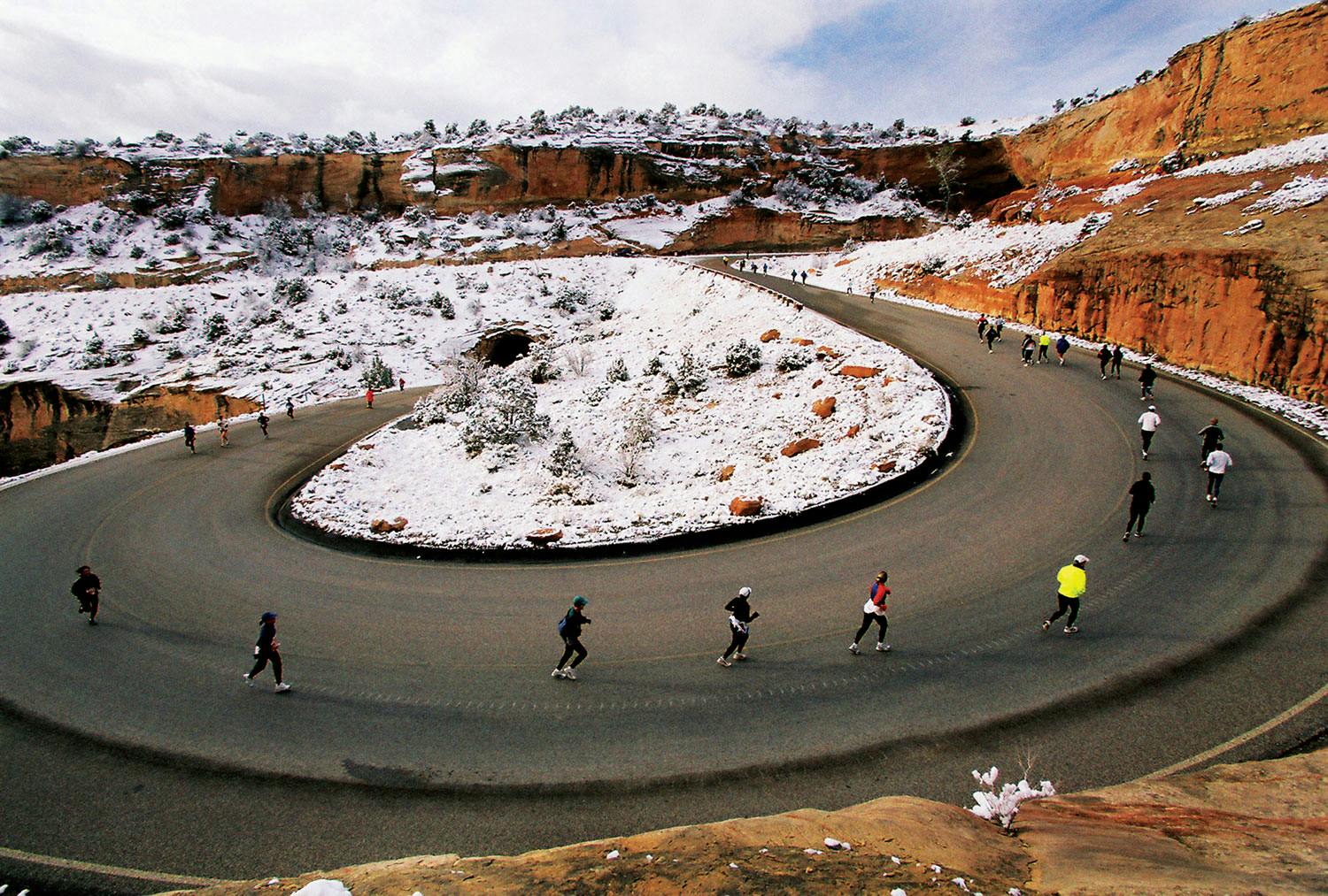 People run up a road switchback. The ground in the middle is covered with snow and the whole area is surrounded by sheer red rock walls. 