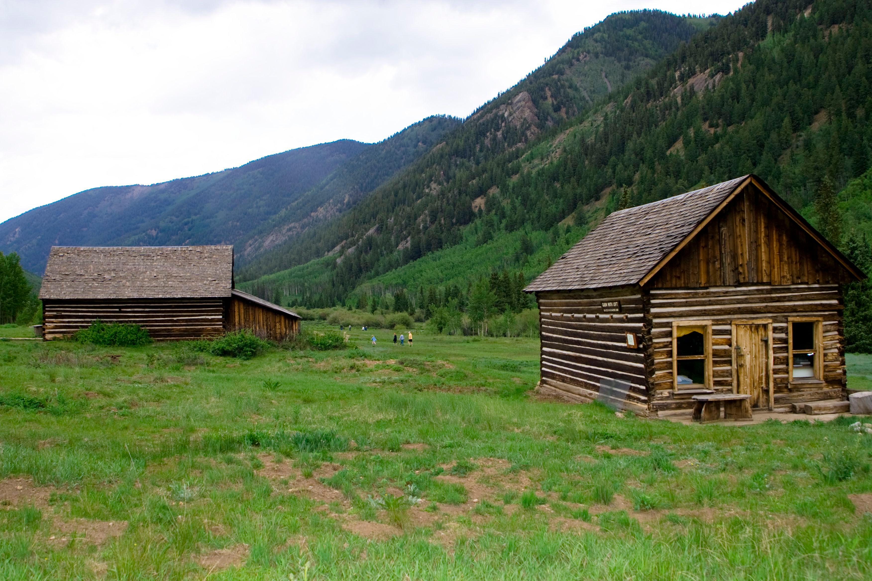 Two log cabins stand in a green field at the base of a steep, forested mountainside in the Ashcroft ghost town.