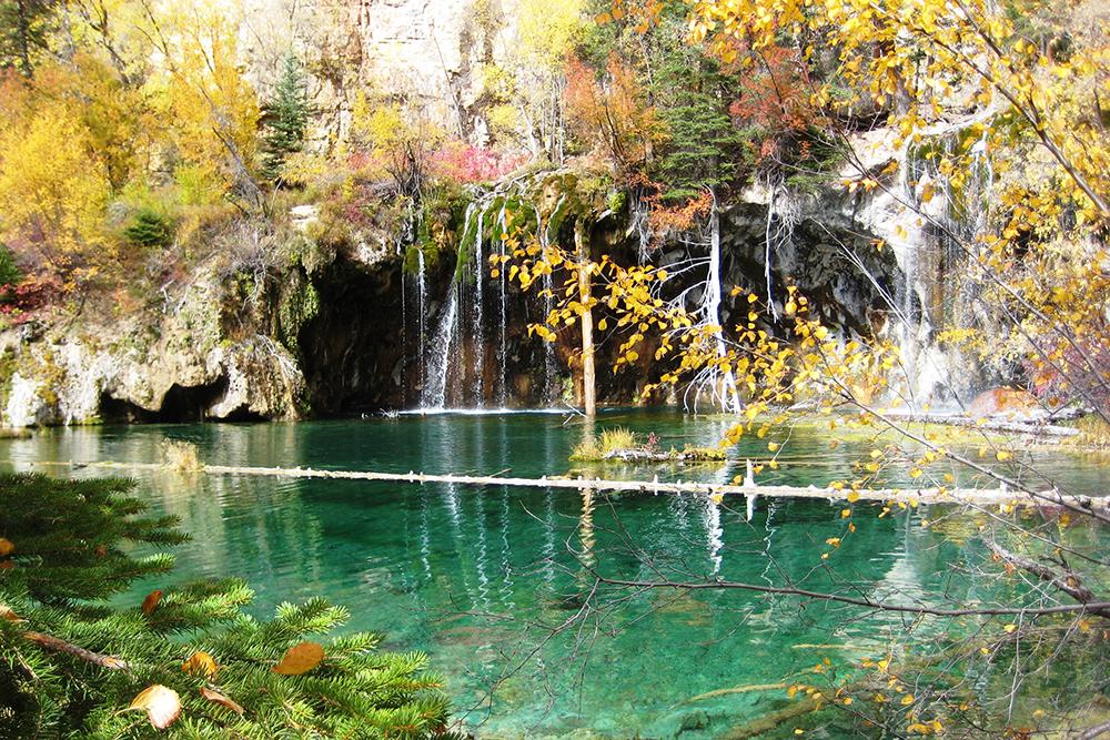 The teal, glassy surface of Hanging Lake is surrounded by fall foliage in hues of green, gold and red. 