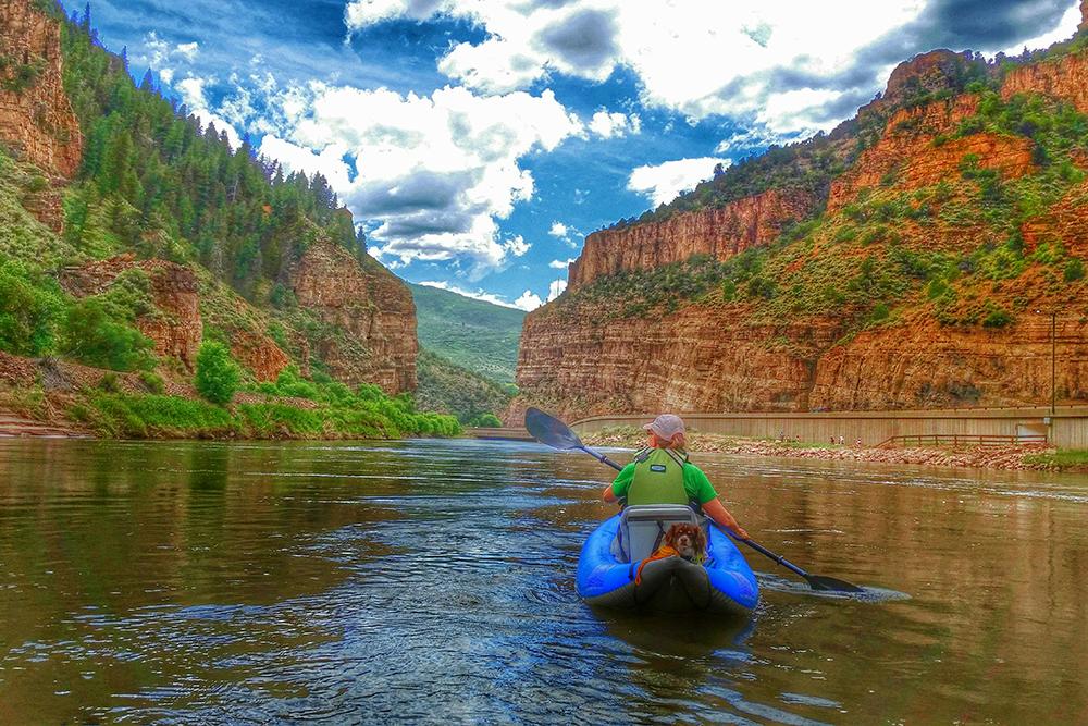 A blue kayak with a green-t-shirt-wearing paddler makes its way through a calm Glenwood Canyon. The walls of the canyon are red with sedimentary lines through them. The walls are dotted with evergreen trees and greenery.