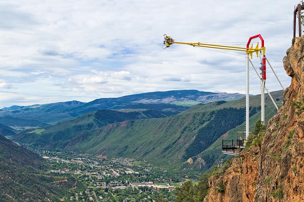 Green mountains meet a white sky above the town of Glenwood Springs. On the right the Giant Swing at Glenwood Caverns Adventure Park is yellow and red with a group of people perpendicular to the ground on the swing.