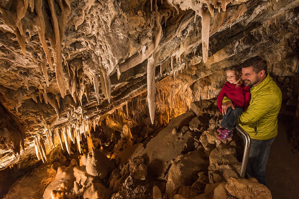 A father in a green jacket holds a toddler in a red jacket looking out at stalactites in Glenwood Caverns.