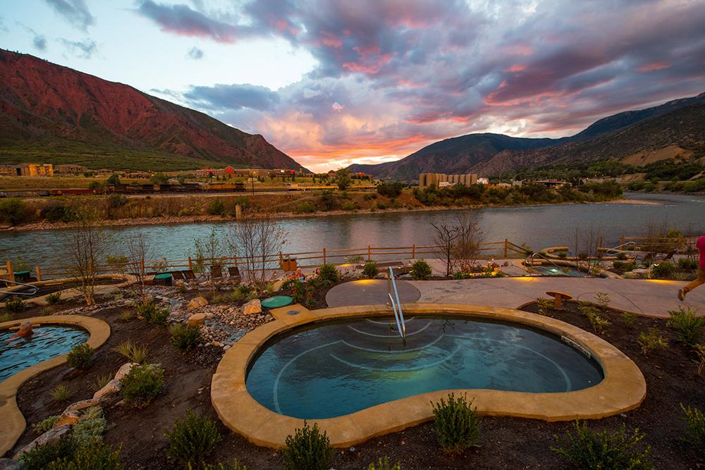 A kidney-bean shaped teal pool sits near other pools at Iron Mountain Hot Spring on the banks of the river. In the distance are the buildings of town in Glenwood Springs. The sun is setting behind red-walled mountains in the distance. 