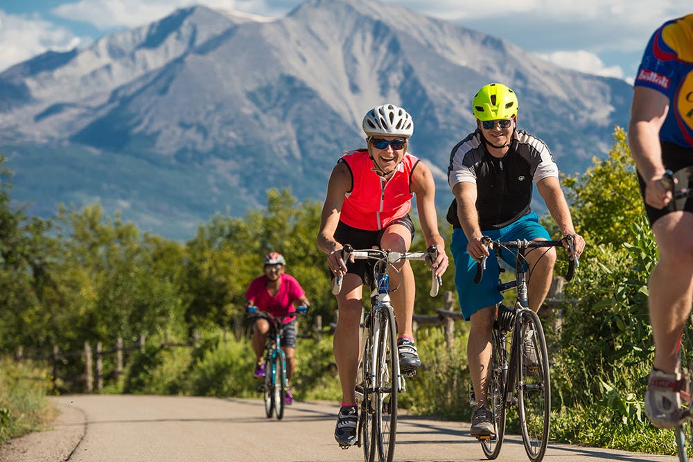 Three people, two wearing bright red tops and one wearing a black and white top road bike on the Rio Grande Trail. The road is surrounded by green grass and trees and in the distance a massive mountain reaches for the sky. 