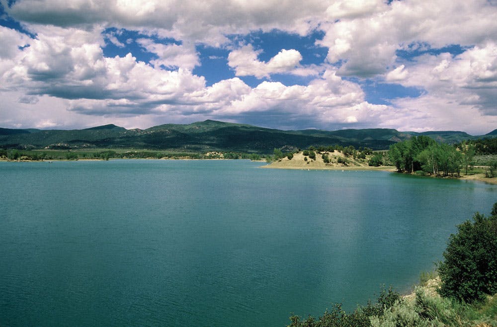 A bright, teal-colored lake sits peacefully beneath a blue sky with flatbottomed clouds floating by