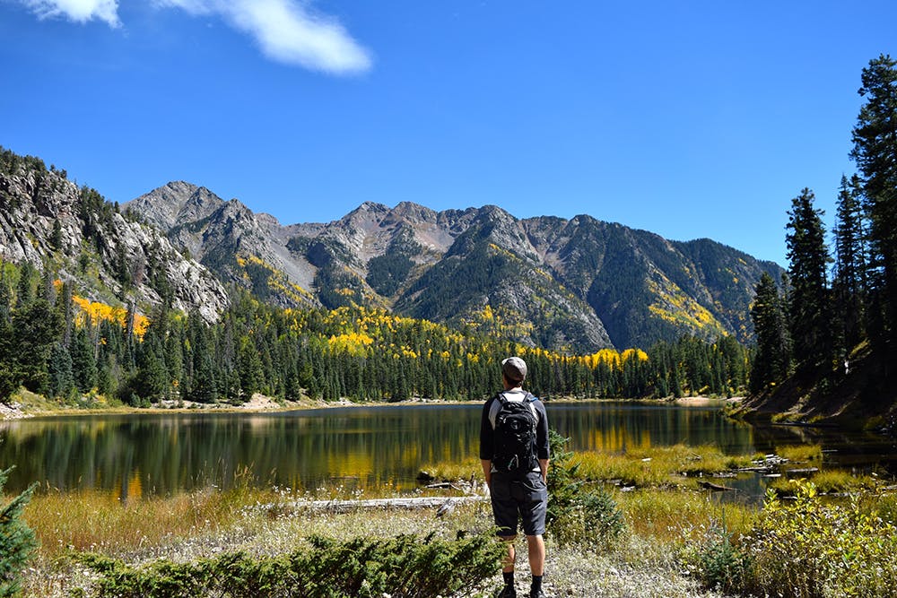 A hiker wearing a backpack looks out on a mountain lake. The water is surrounded by evergreen trees that are dotted with golden trees that go up to Rocky Mountain peaks and a blue sky.