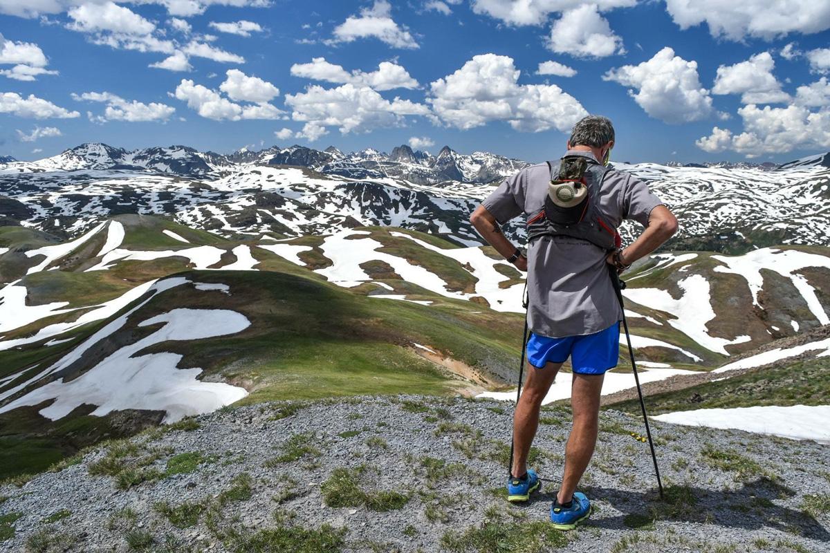 A person holding hiking poles looks out at snow-covered patches atop mountain peaks.