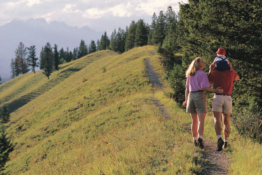 A couple, the woman wearing a lilac-colored shirt with shorts and the man wearing a red-shirt with khaki shorts, walks along a ridge on a dirt path. A child sits on the shoulders of the man wearing a red hat. To the left of the path is a hill of green grass. In the distance evergreen trees meet a hazy sky.