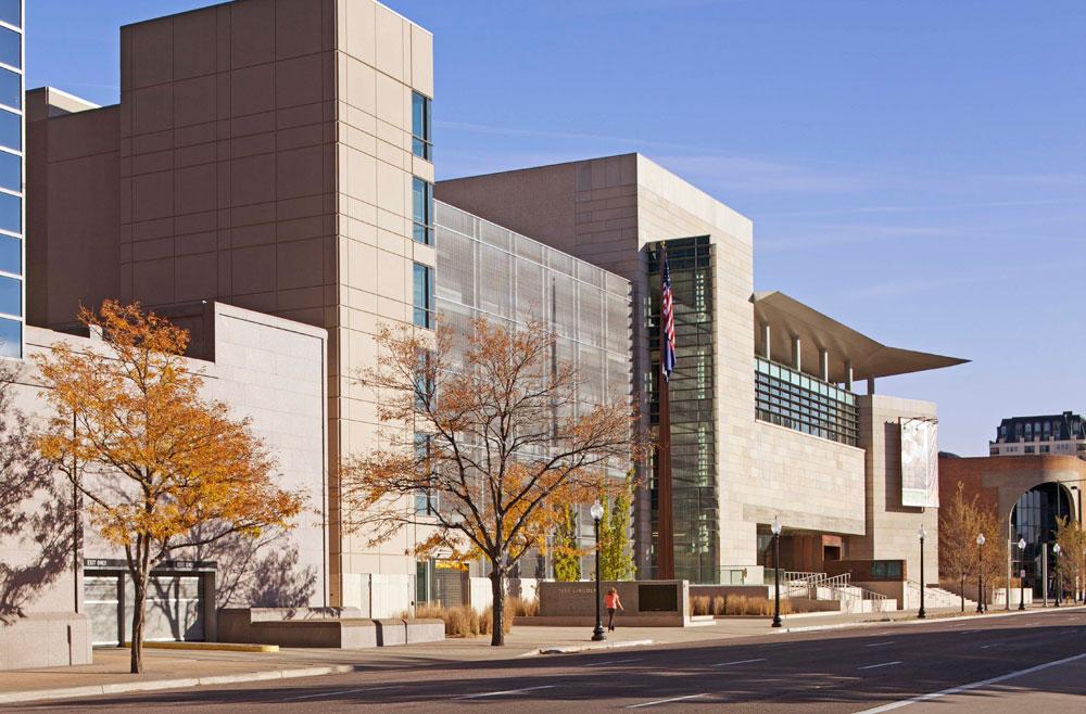 It's fall in Denver, Colorado, and young trees planted along a wide sidewalk have turned orange and have begun to loose their leaves. A tall white-stone and window-faces building stands behind them.