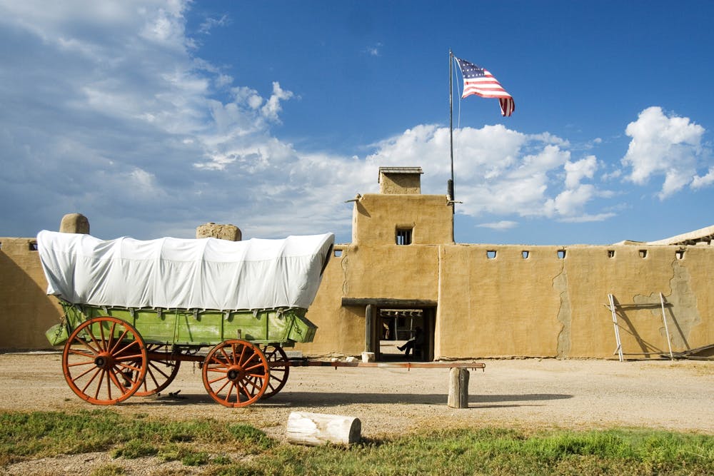 A green wooden wagon with red spoke wheels and a white canvas top rests in front of the Old Fort Historic Site near La Junta.