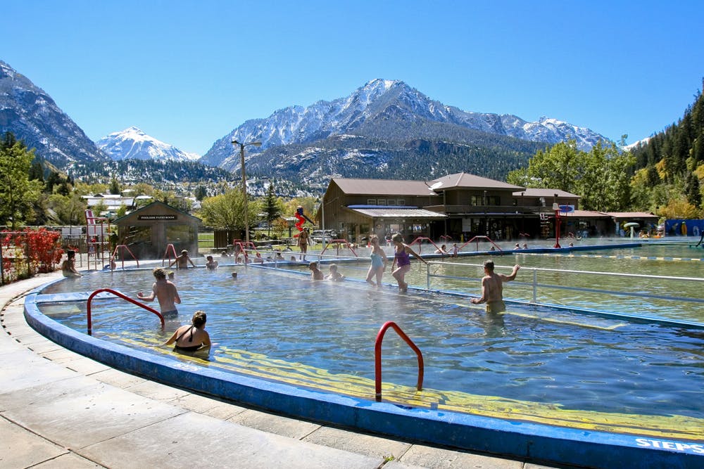 People wade in the steaming Ouray Hot Springs Pool with a backdrop of snow-covered mountains.