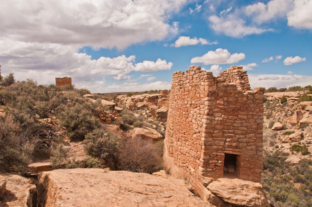 A tall structure made of red and tan clay bricks stands amid large rocks and shrubs in the desert west of Cortez, Colorado.