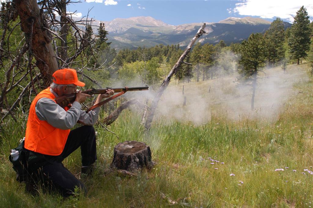 A man wearning a hunters-orange vest and hat kneels with his muzzleloader and points it off into the distance