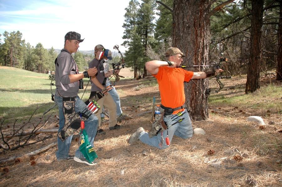 One man in hunters orange kneels and pulls back the arrow on his bow, behind him, two others carrying lots of archery gear wait their turn