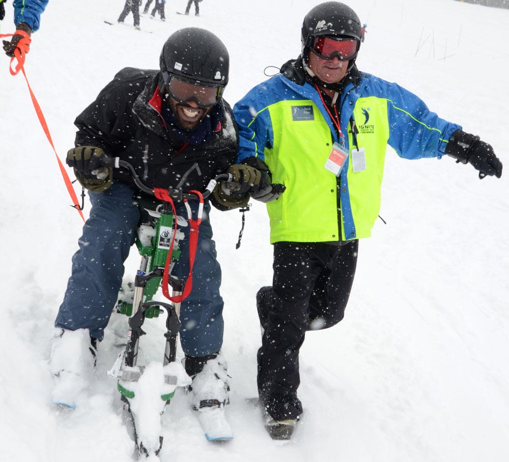 An instructor in a blue and neon-green jacket guides a new student in a black jacket through the snow on a ski bike in Colorado.