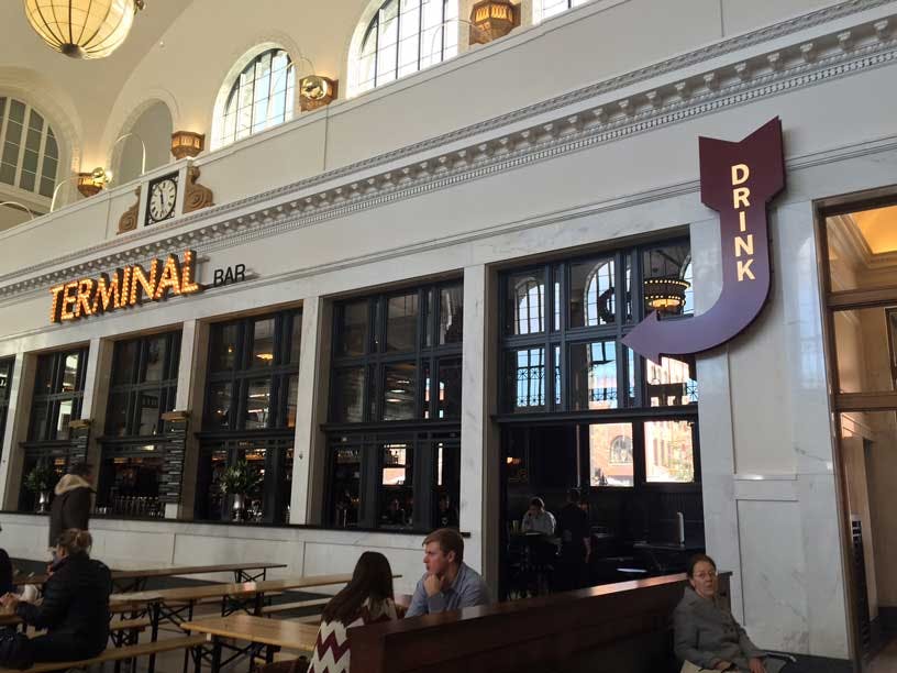 People sit at picnic-style tables outside a bar in Union Station in Denver, Colorado. The bar name "Terminal" is lit with a marque-style  sign.