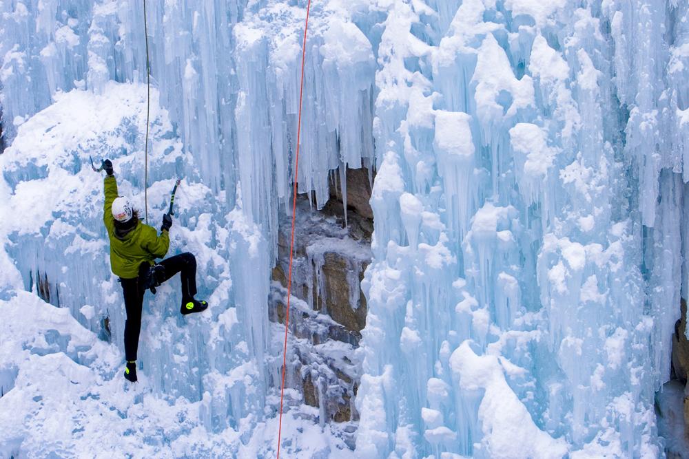 An ice climber in a green jacket with black pants climbs up bright-white ice structures.