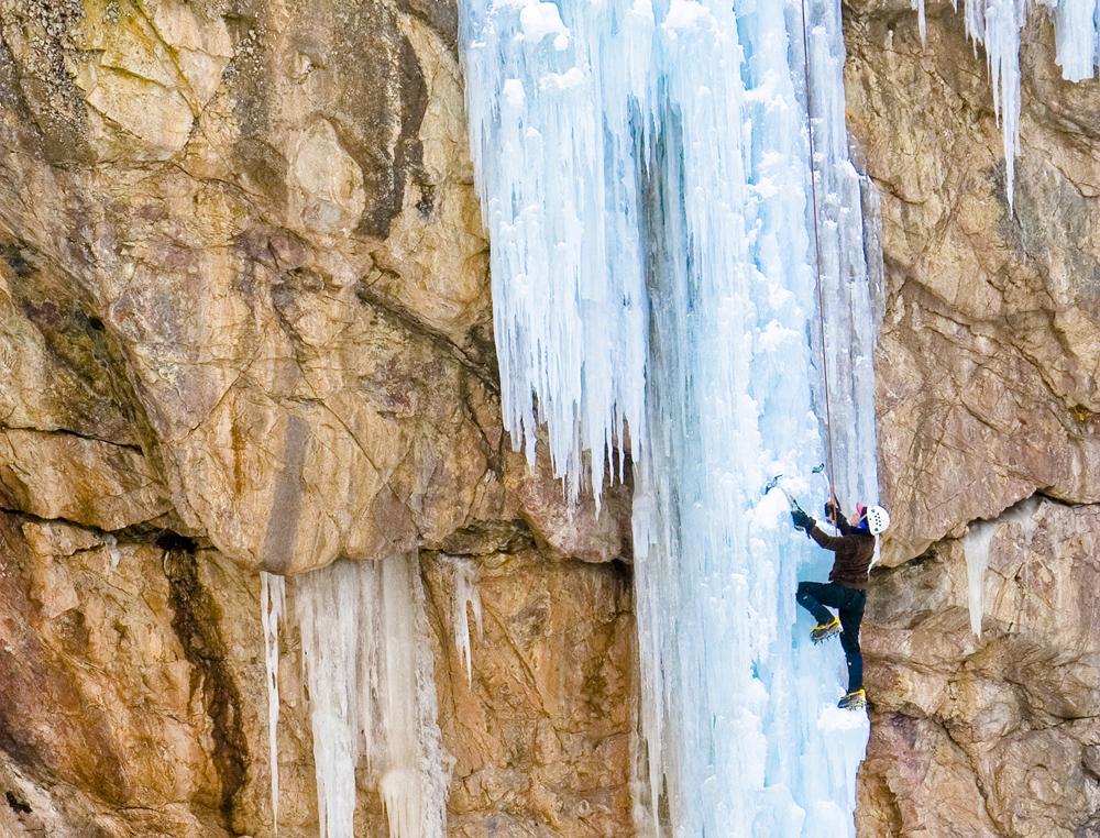 A solo ice climber makes their way up bright-white ice sculptures on a sand-colored rock face.