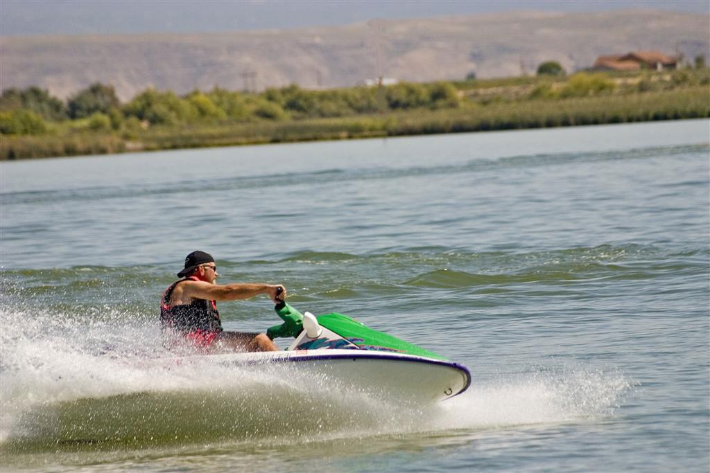 A man on a green jet ski with a backwards black baseball cap jet skis on a greenish blue Sweitzer Lake. In the back there is a red-roofed house with green bushes along the lake's shores.