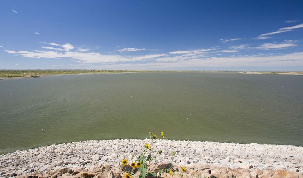 The rocky shore of John Martin Reservoir. In front of the shore is the green water of the body with a blue sky and white clouds.