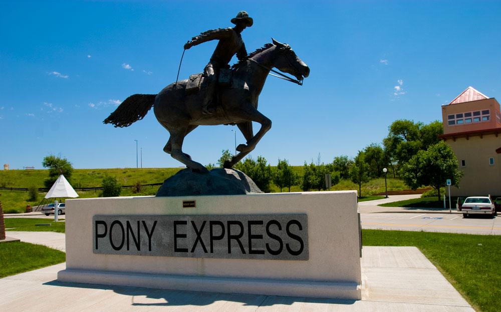 In Julesburg, Colorado, a statue of a person riding a galloping horse commemorates the Pony Express Route. Behind the statue is a green hill and a blue sky.