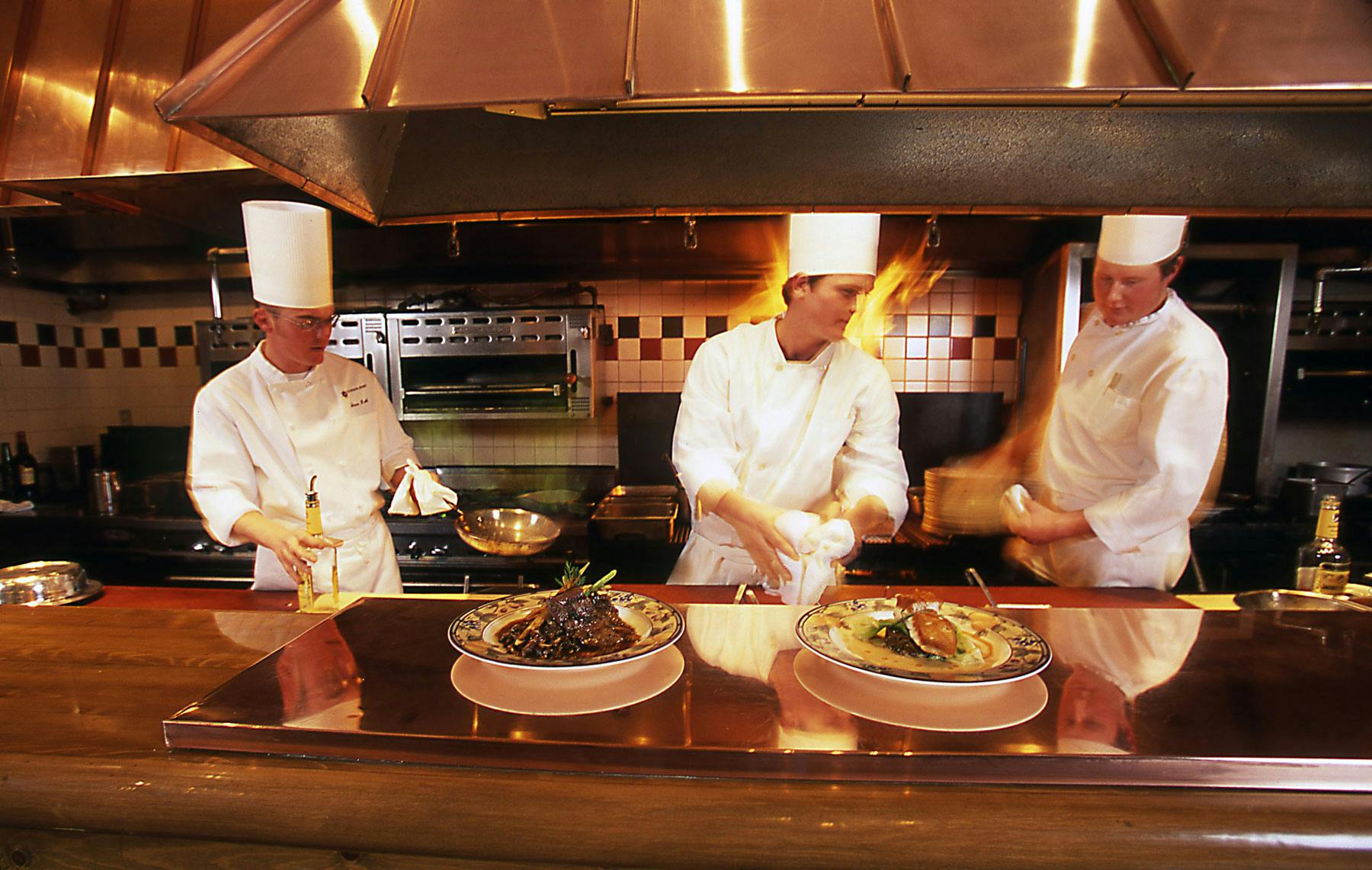 Three chefs do their thing in a kitchen behind a copper hood and bar, where two plates are lined up to be taken to their table