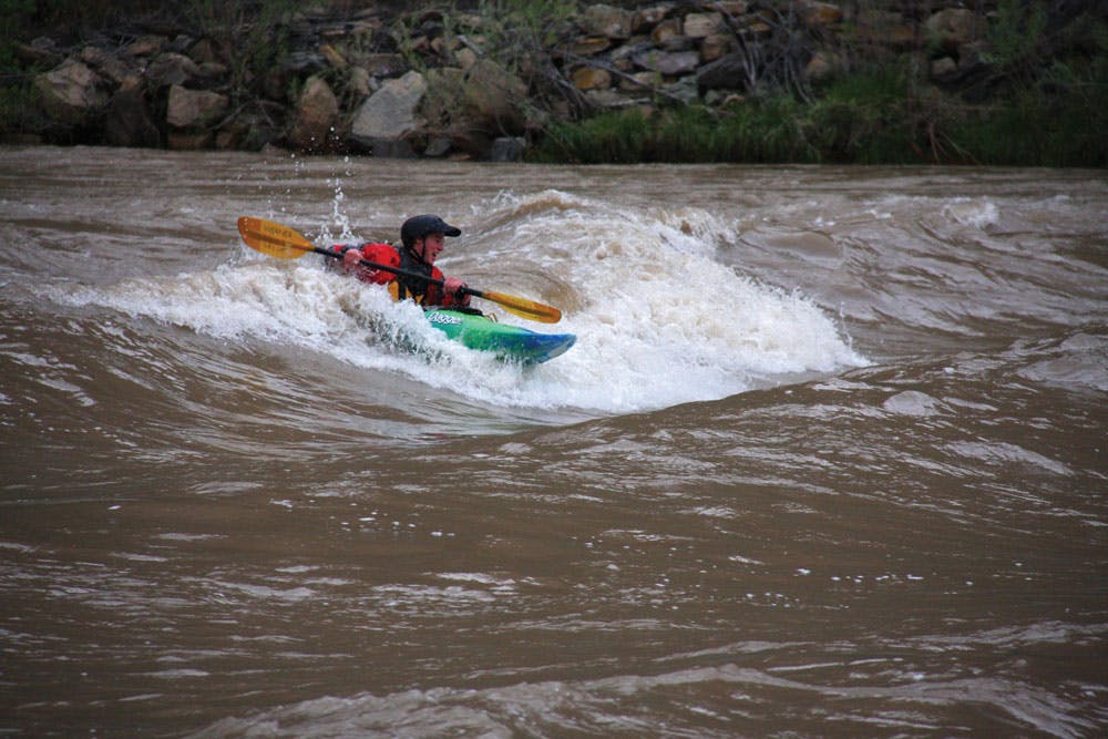 A whitewater kayaker in a red jacket sits in a green and blue kayak in the middle of a white wave in the midst of a brown river. In the background there's a rocky shoreline.