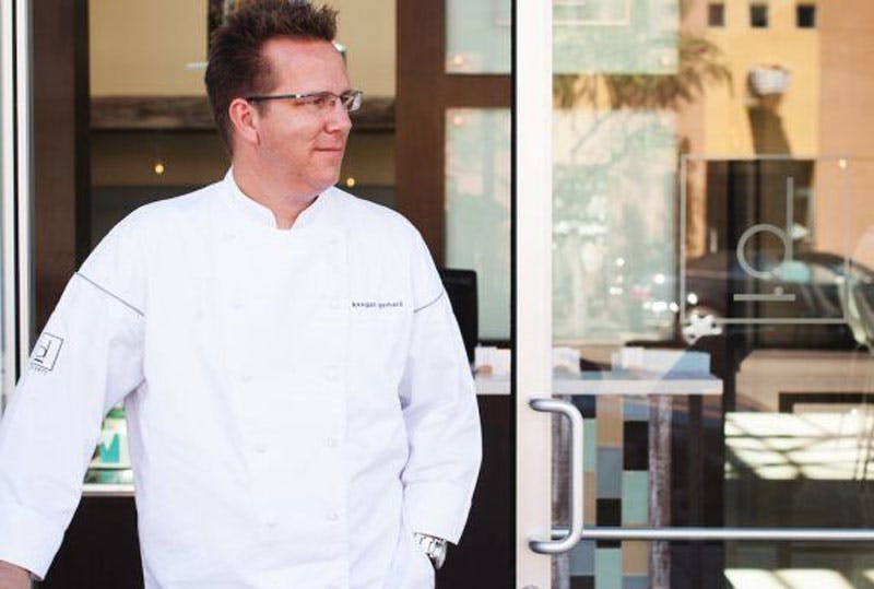 In Denver, Colorado, chef Keegan Gerhard stands in a white chef coat and rectangular-framed glasses and looks off in the distance outside D Bar.