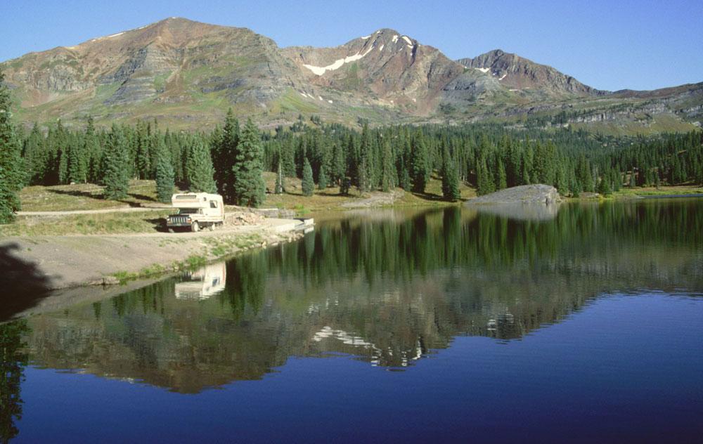 An RV sets up camp on the reflective Lake Irwin near Crested Butte, Colorado. The mountains and blue sky stand above the lake and reflections shimmer in the water.