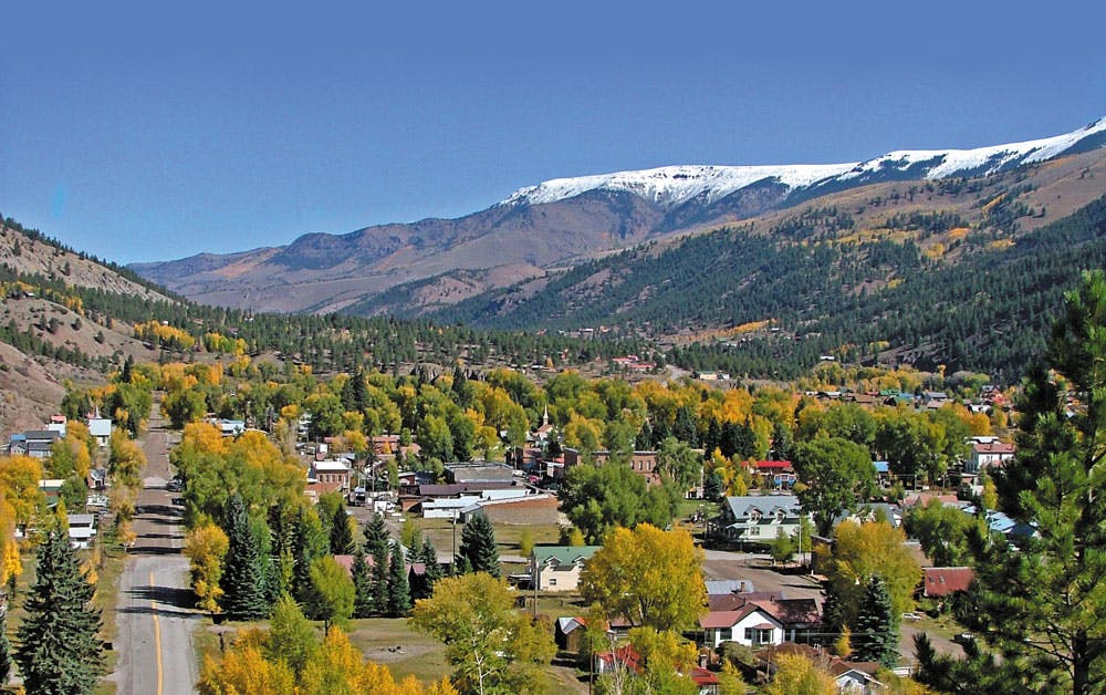 An aerial view of the small town of Lake City show streets lined with trees and homes on a sunny day; snow sits atop a mountain ridge