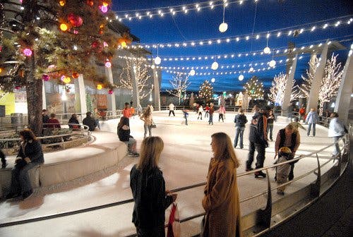 A nighttime shot of the ice rink at Belmar in Lakewood with ice skaters moving around. There are twinkle lights hung up across the air above the rink.