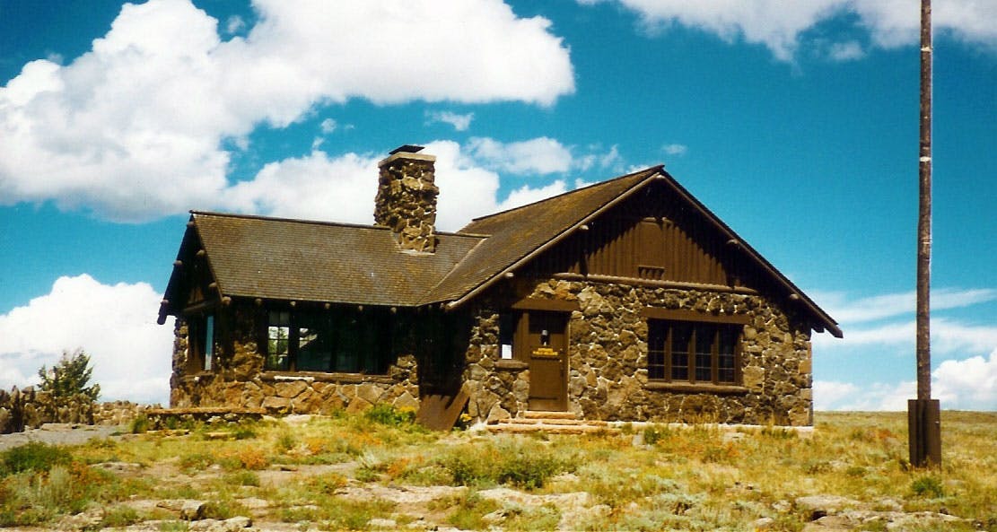 A brick and brown-wood-trimmed building with a chimney sits in a field of grass